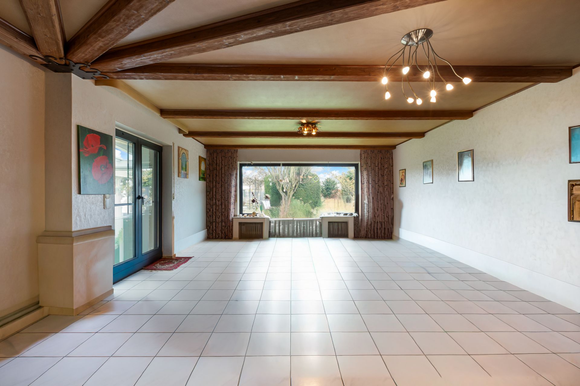 Helles Wohnzimmer mit weißen Fliesen, Holzbalken an der Decke und großem Fenster mit Blick nach draußen.