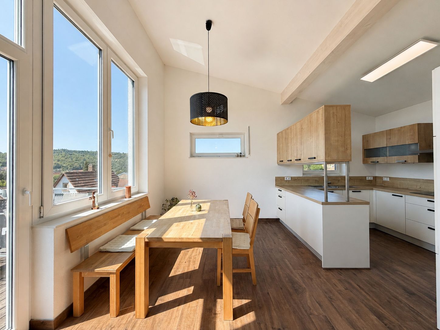Helle Küche mit Holztisch, Bank und Stühlen sowie großen Fenstern und Einbauschränken.