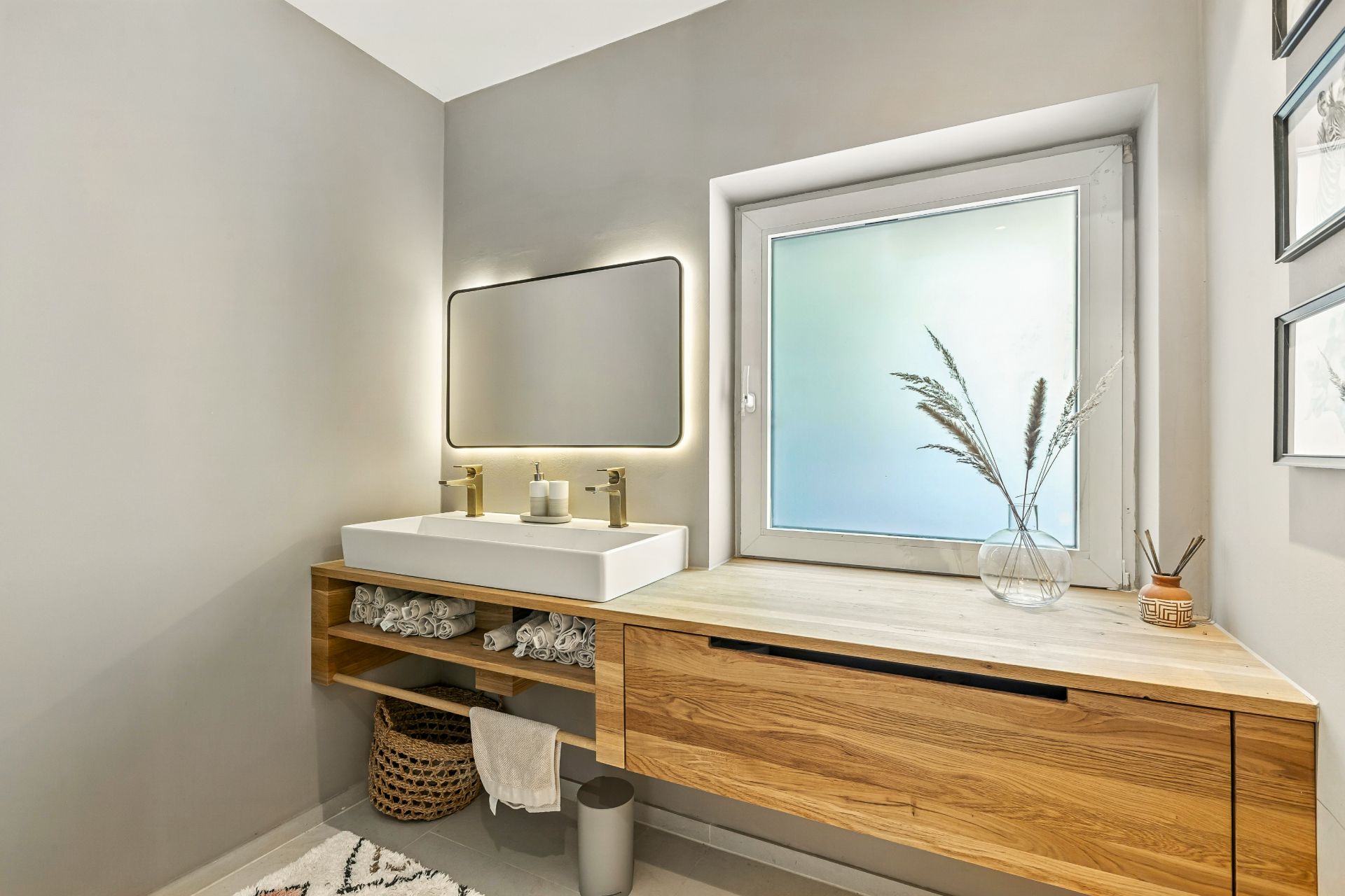Modernes Badezimmer mit großem Waschbecken, Holzschrank und beleuchtetem Spiegel neben einem Fenster.