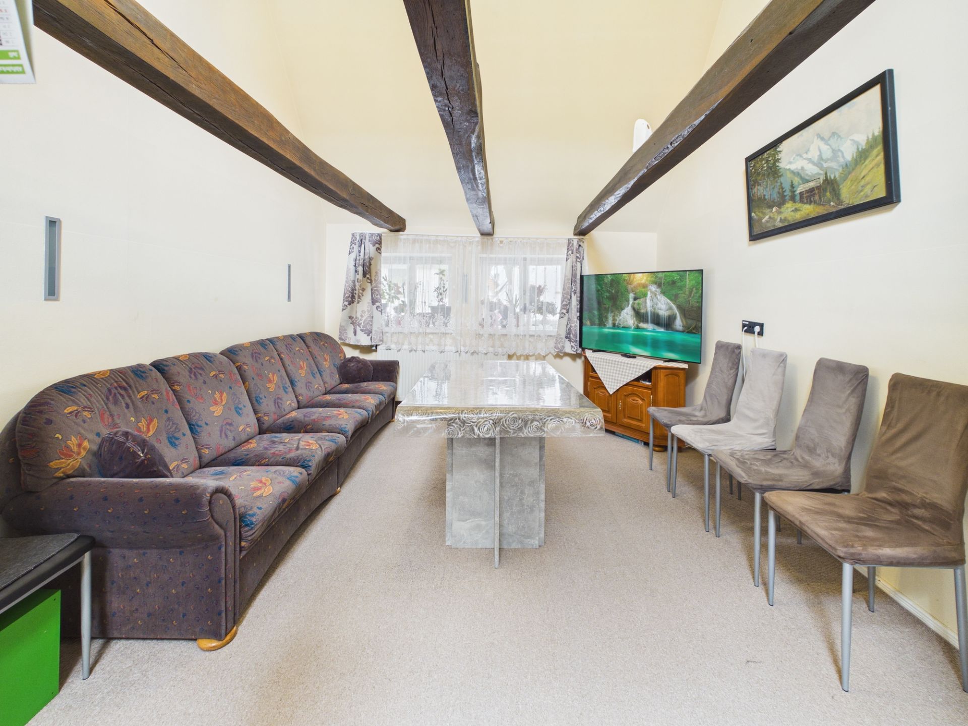 Wohnzimmer mit Sofa, vier Stühlen, Tisch, Fernseher und sichtbaren Holzbalken an der Decke.