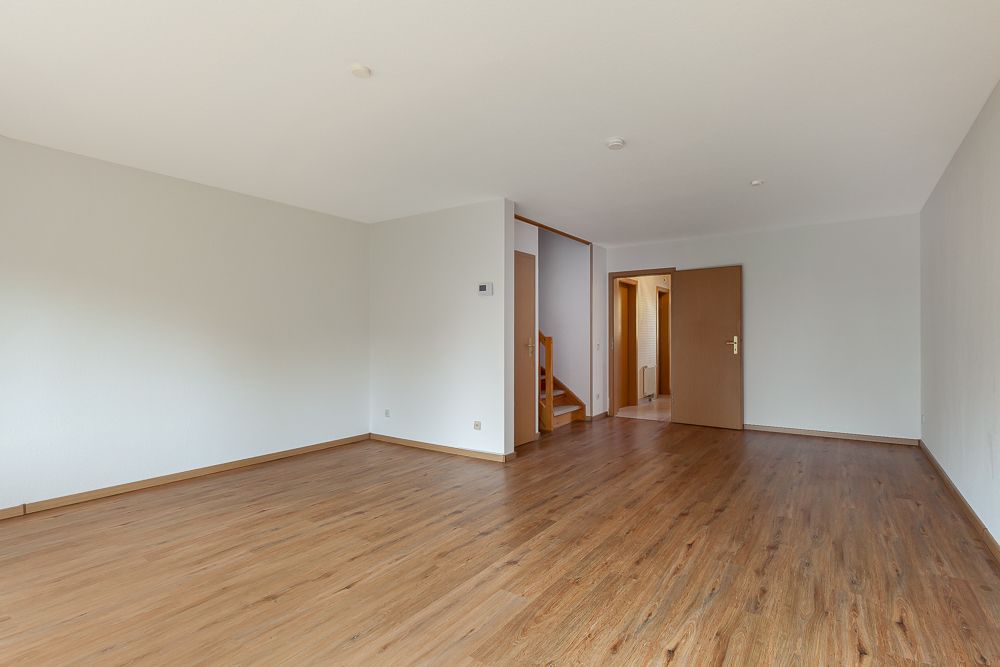 Leeres Wohnzimmer mit Holzfußboden, weißen Wänden und offener Tür zu einem Flur mit Treppe.