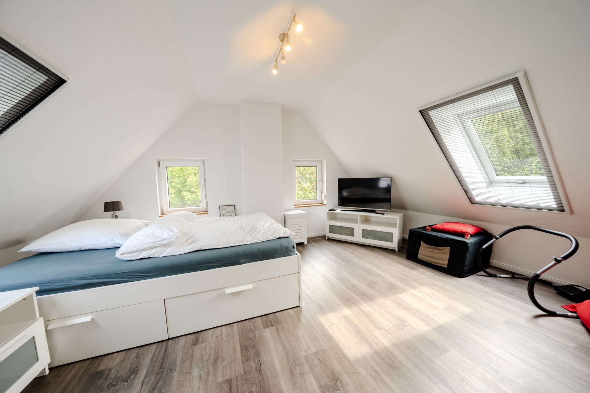 Helles Schlafzimmer mit Doppelbett, Fernseher, zwei Fenstern und schräger Decke.