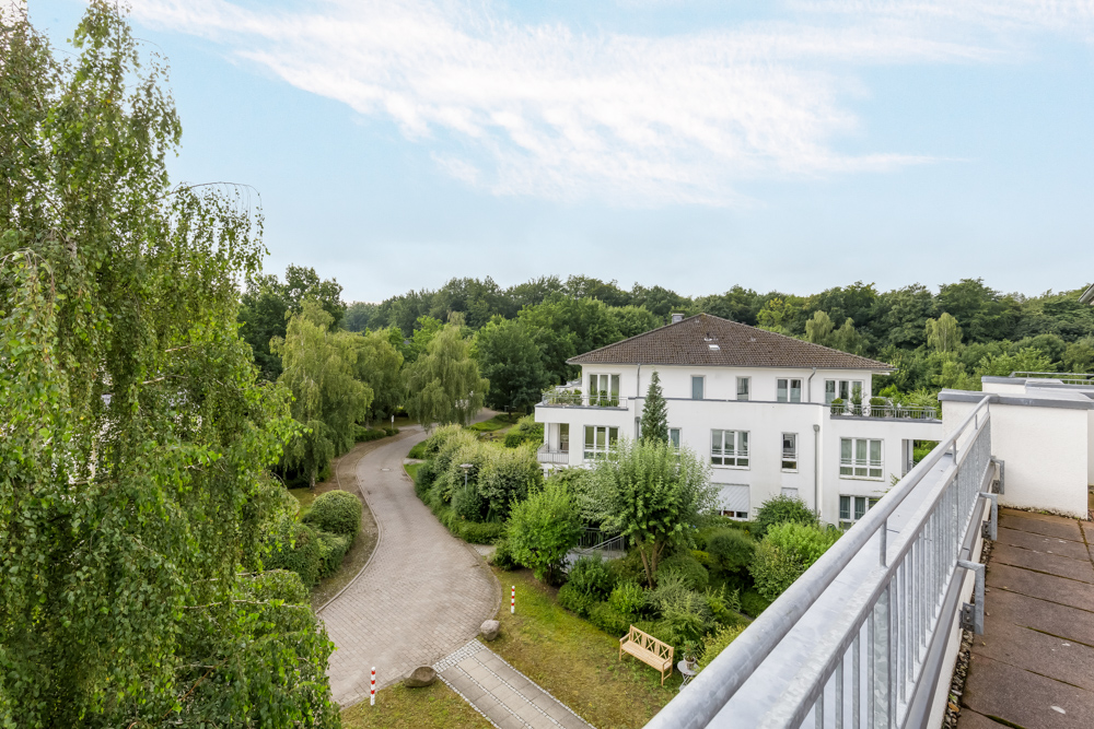 Blick von einem Balkon auf ein weißes Wohnhaus und eine begrünte Einfahrt.