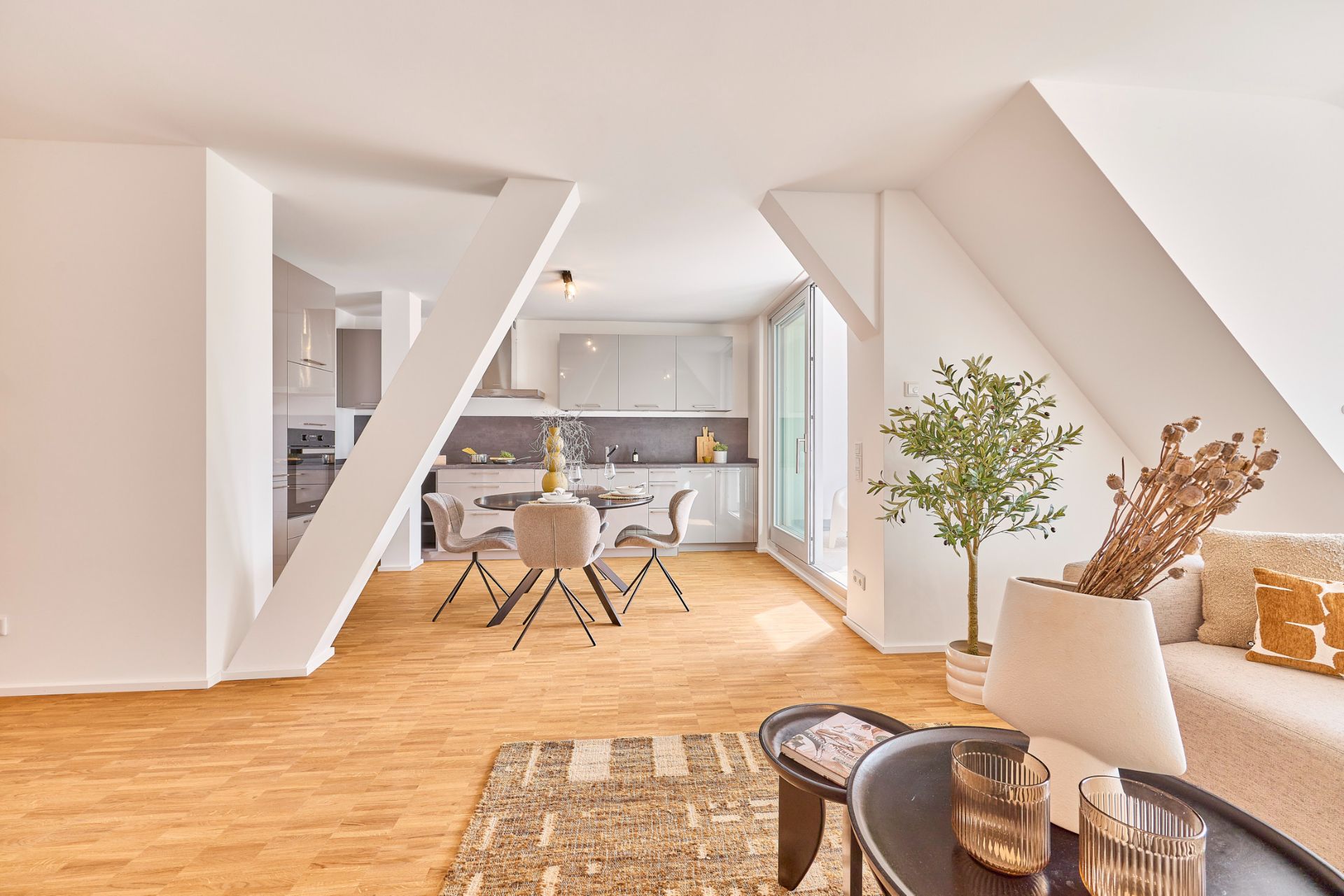 Helles Wohnzimmer mit offener Küche, Esstisch, Sessel, Teppich und Zimmerpflanzen.