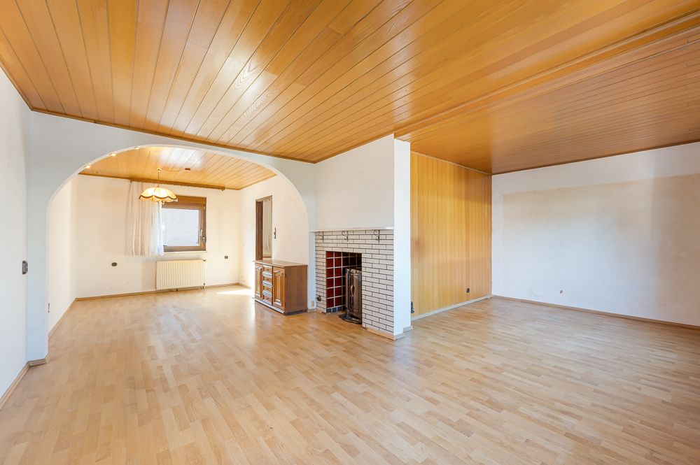 Leeres Wohnzimmer mit Holzboden, Holzdecke, Kamin und Durchgang zum Essbereich.