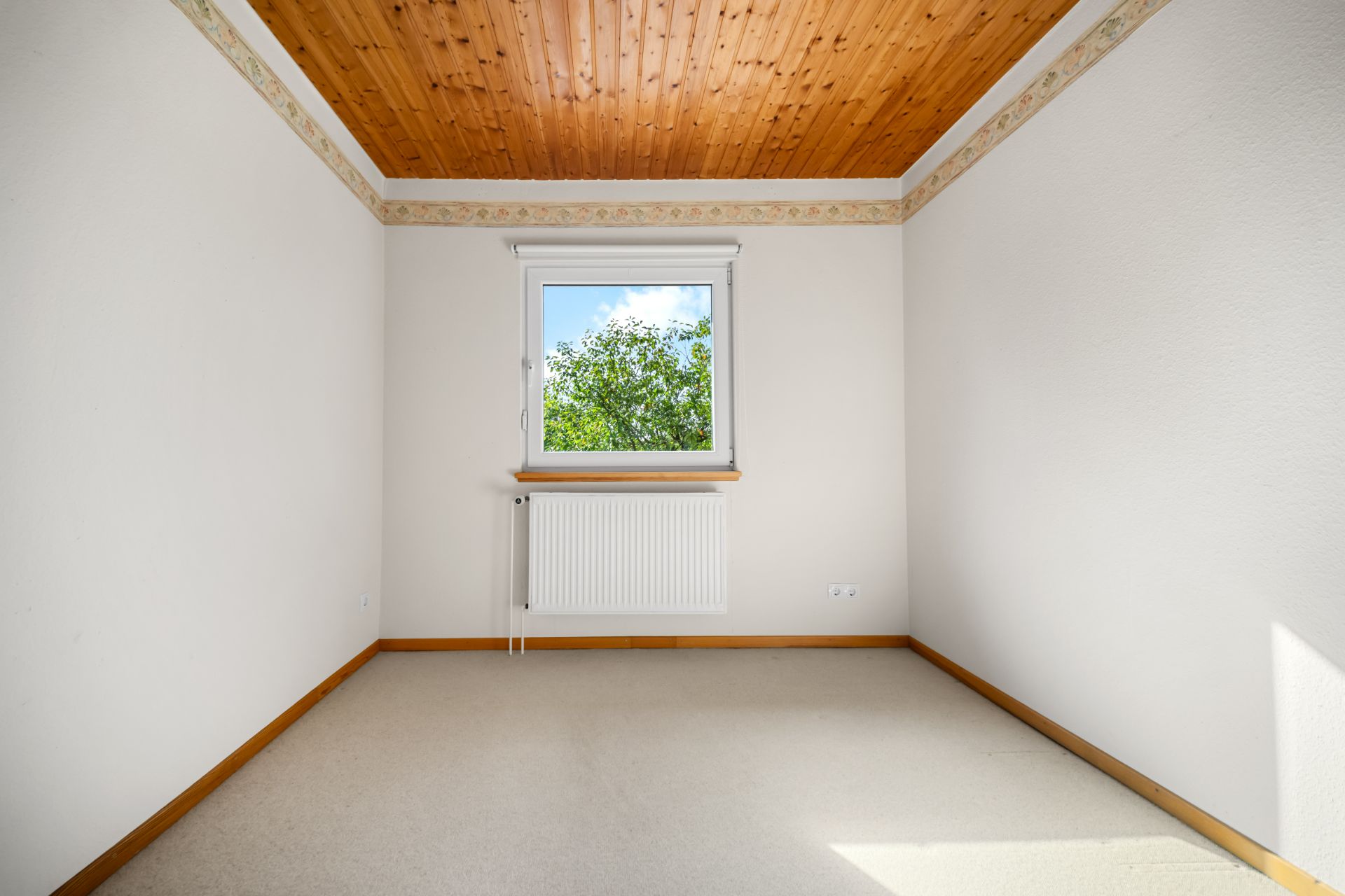 Leeres Zimmer mit Holzdecke, Teppichboden und einem Fenster mit Blick auf grüne Bäume.