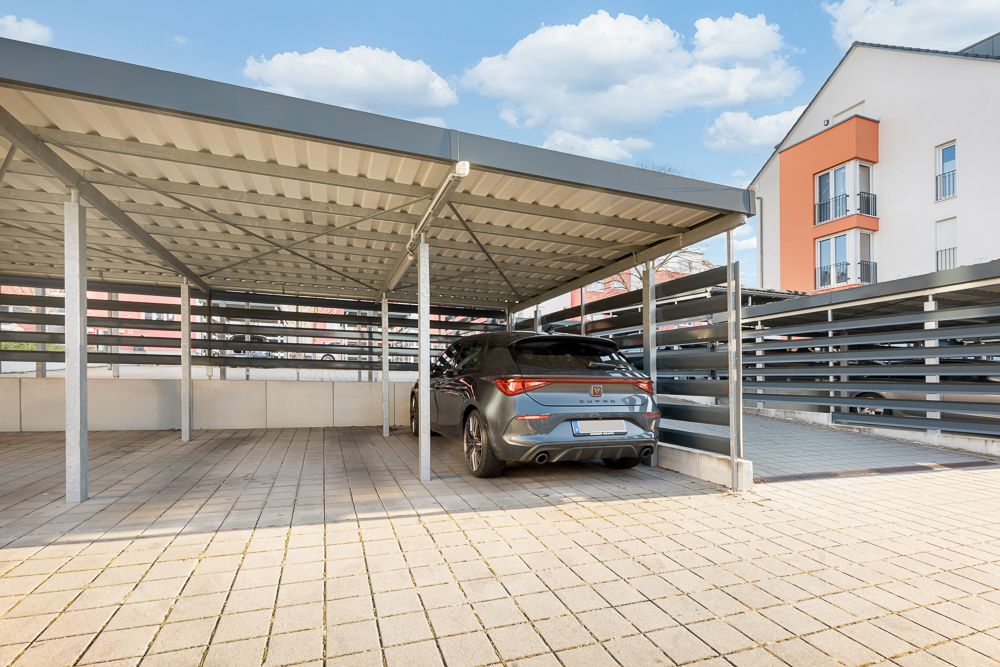 Überdachter Stellplatz mit einem geparkten Auto auf gepflastertem Boden neben Wohngebäuden.
