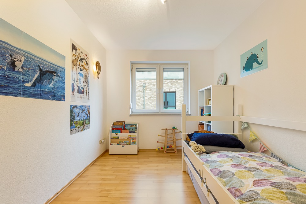 Helles Kinderzimmer mit Einzelbett, Spielzeug, Bücherregal und bunten Wandbildern.