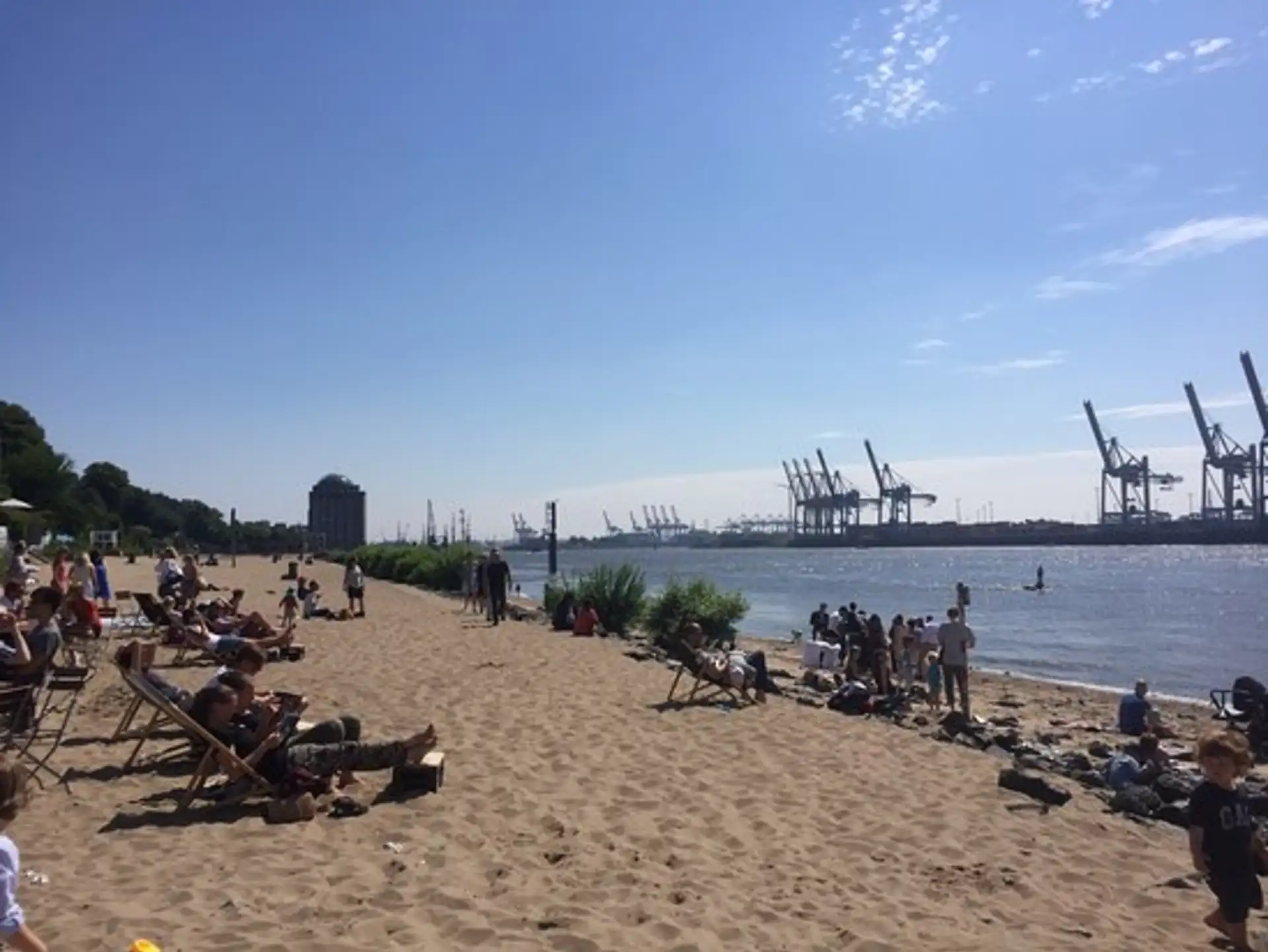 Menschen entspannen am Sandstrand an einem Fluss mit Hafenkränen im Hintergrund.