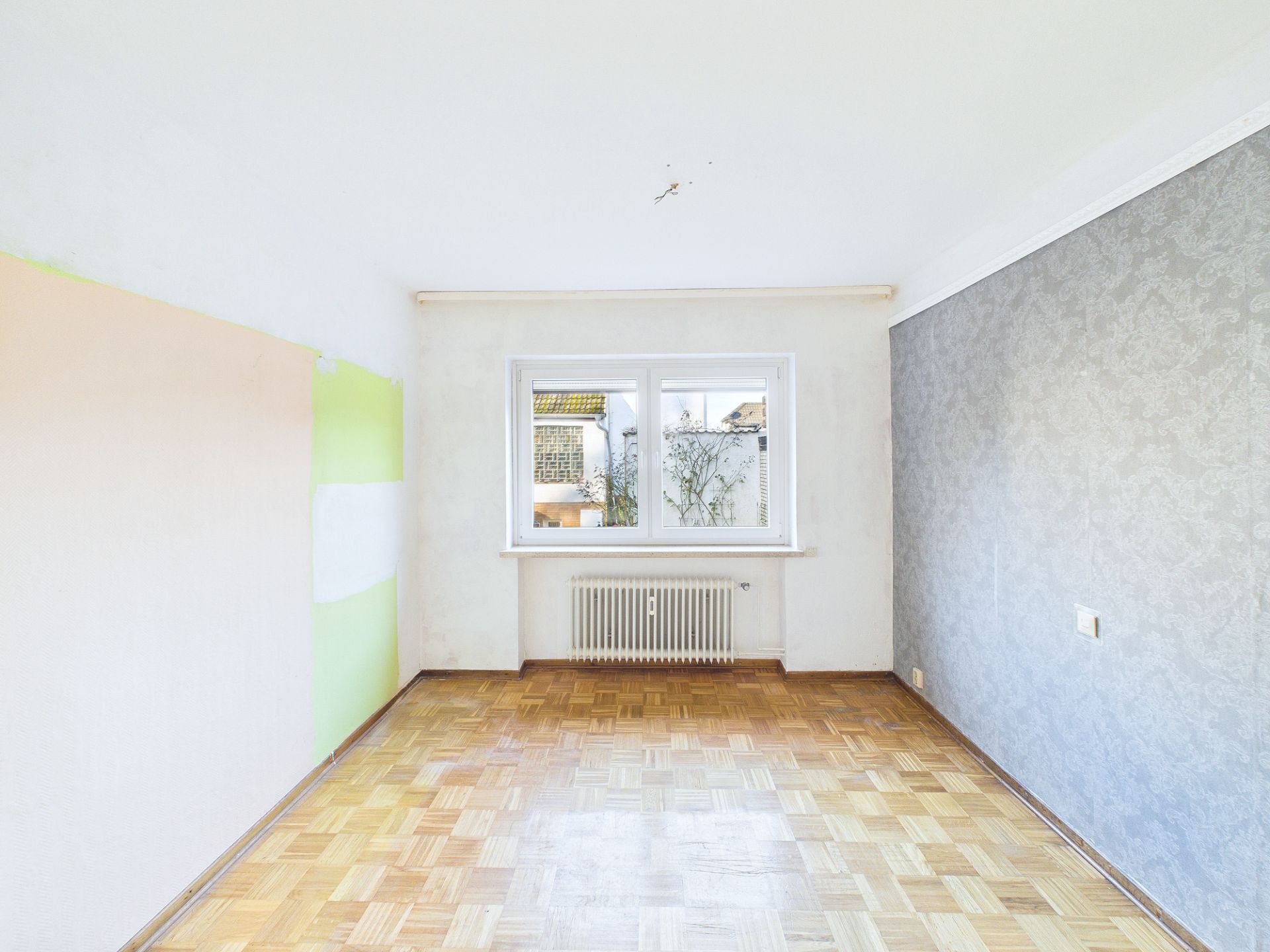 Leeres Zimmer mit Parkettboden, Fenster und Heizkörper, eine Wand teils grün gestrichen.