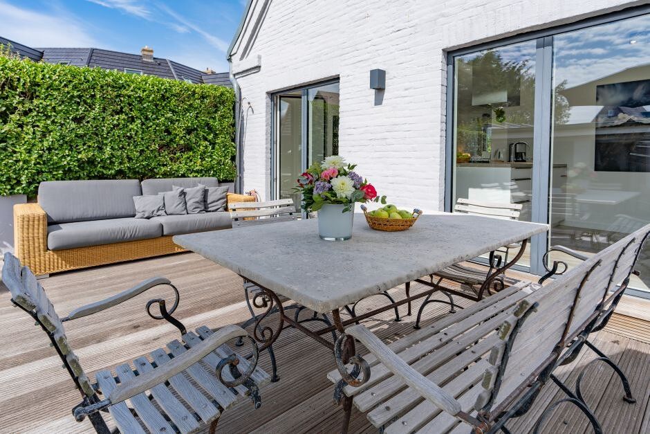 Holzterrasse mit einem Esstisch, Stühlen, Sofa und Blumenvase neben einem weißen Haus.