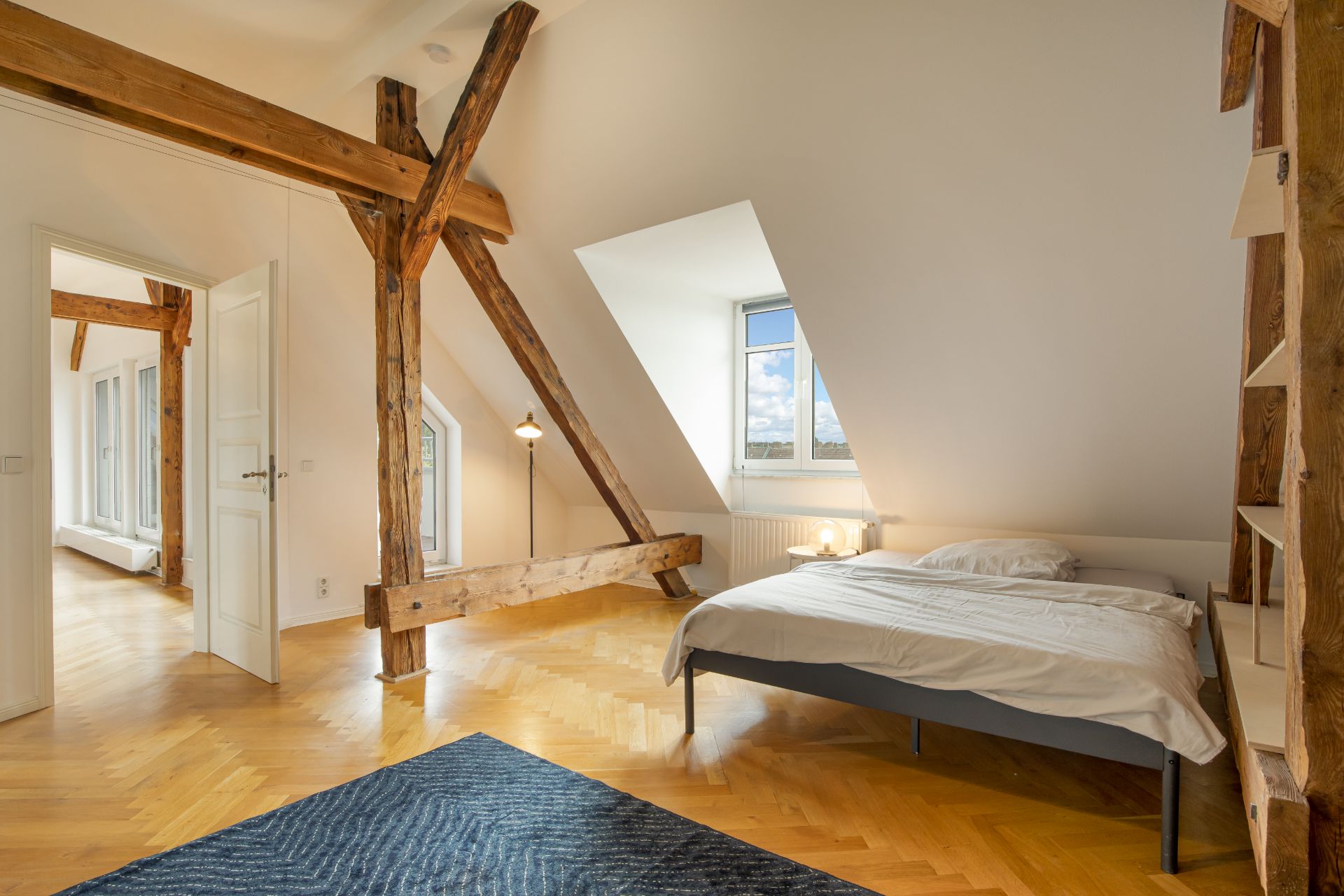 Helles Schlafzimmer mit Holzbalken, Bett, Teppich und großem Fenster.