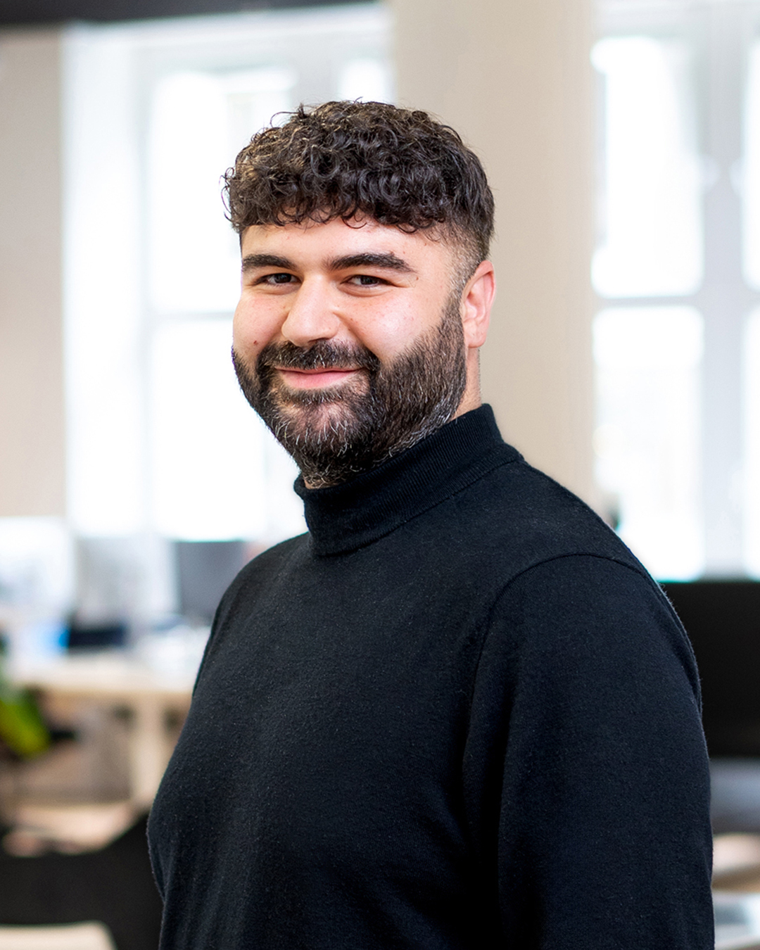 Ein Mann mit Bart und lockigem Haar steht in einem hellen Raum mit Fenstern im Hintergrund.