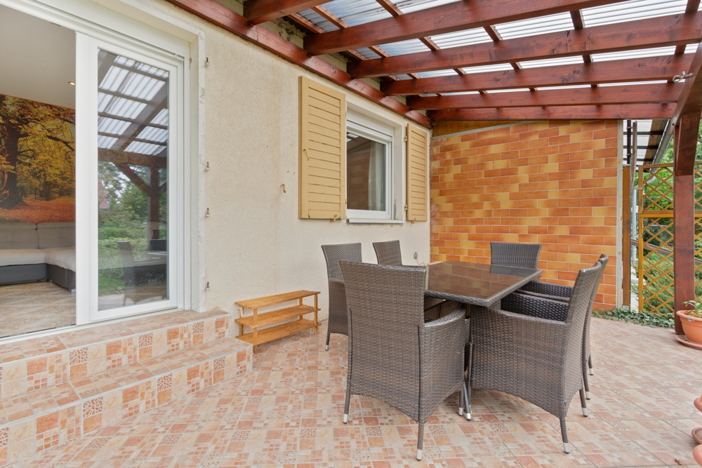Überdachte Terrasse mit Esstisch und sechs Stühlen, Fliesenboden und Zugang zu einem Raum drinnen.