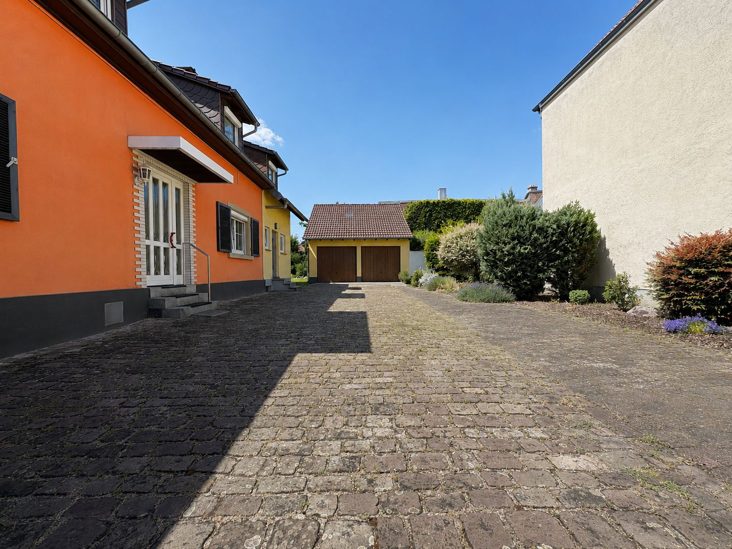 Gepflasterter Hof mit orangefarbenem Haus links, Garage im Hintergrund und Büschen rechts.