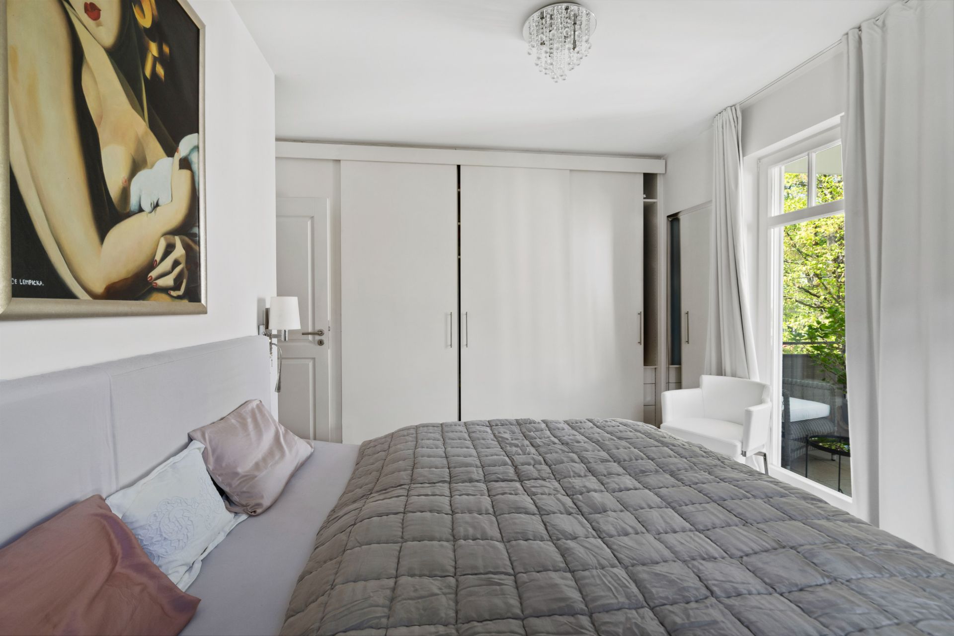 Schlafzimmer mit grauer Bettdecke, weißem Schrank, Fenster und einem Gemälde an der Wand.
