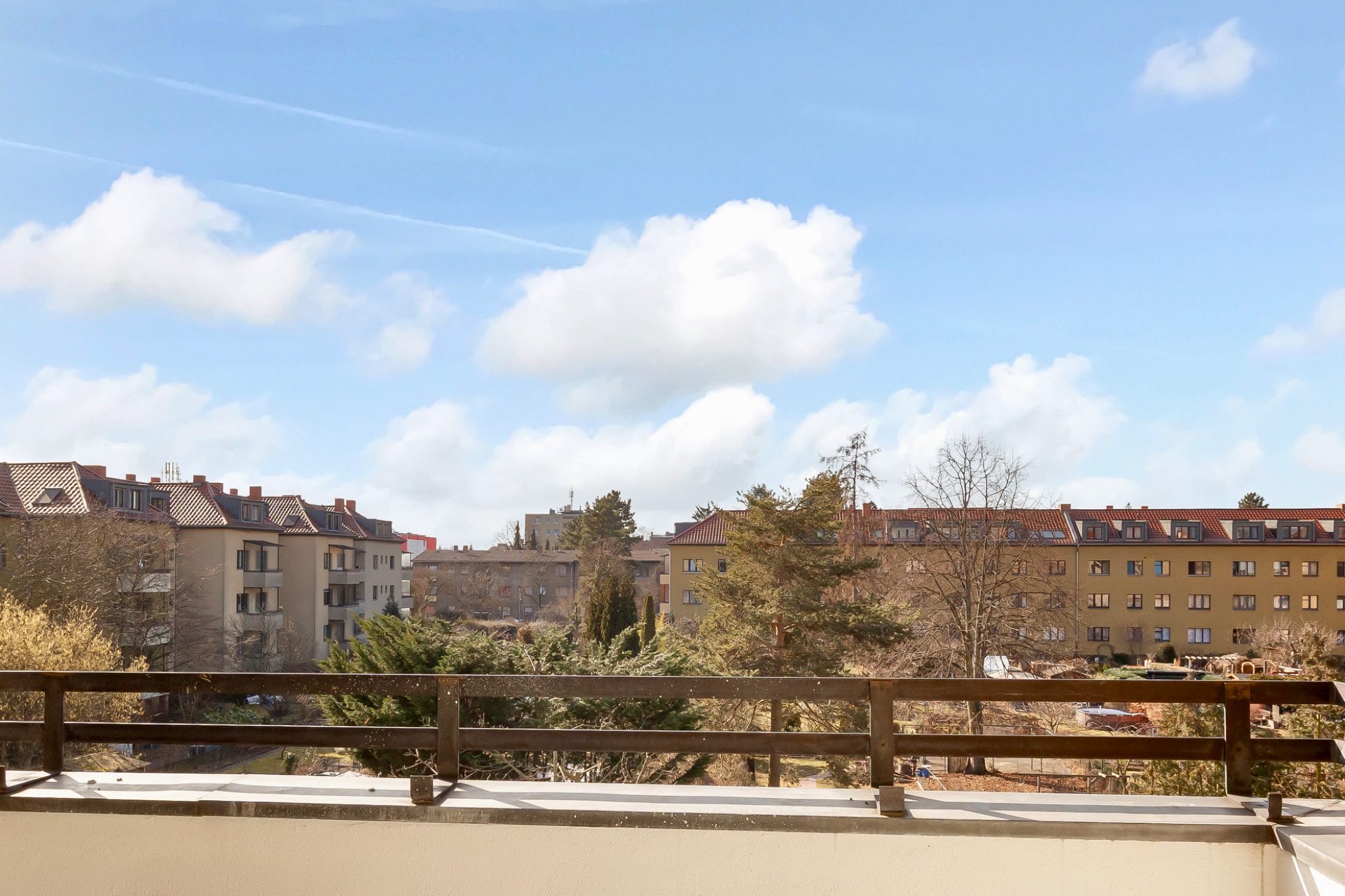 Blick von einem Balkon auf Wohngebäude, Bäume und einen Innenhof bei sonnigem Wetter.