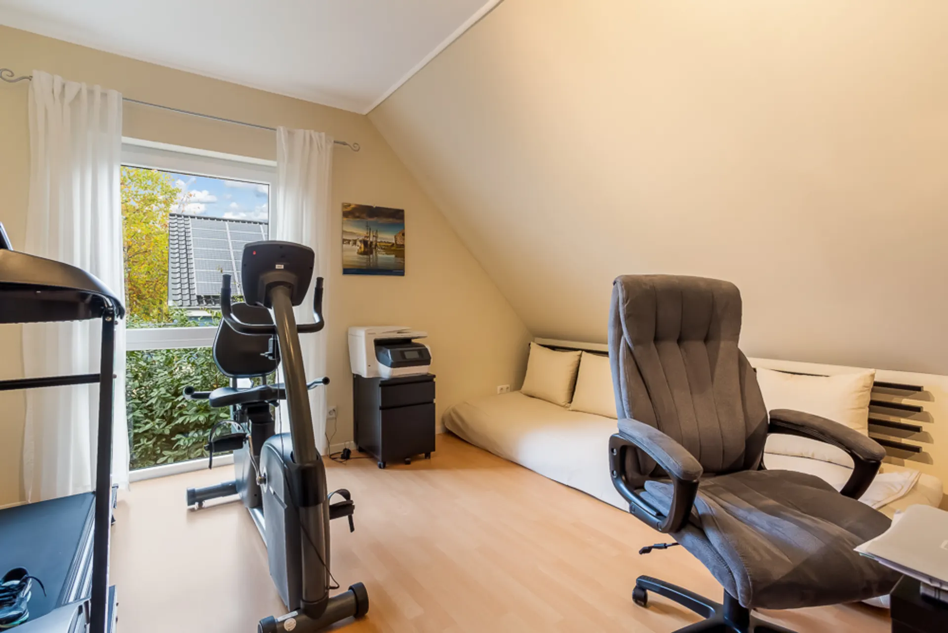 Helles Zimmer mit Heimtrainer, Bürostuhl, Tagesbett und großem Fenster mit Blick nach draußen.
