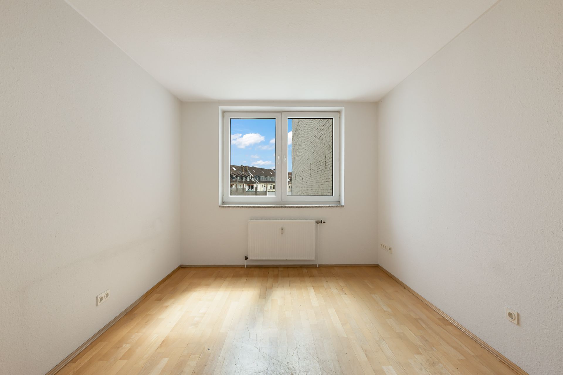 Leerer Raum mit hellem Holzboden, weiß gestrichenen Wänden und einem Fenster mit Blick nach draußen.