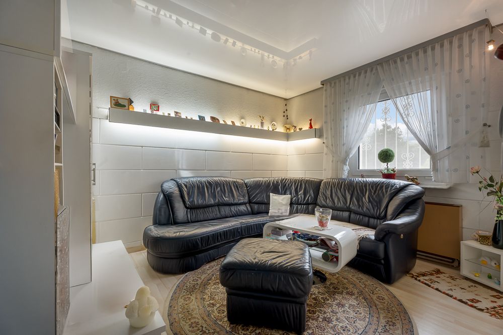 Wohnzimmer mit schwarzem Ecksofa, rundem Teppich, Couchtisch und großem Fenster mit Gardinen.