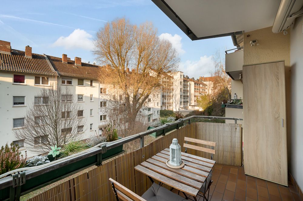 Balkon mit Holztisch, zwei Stühlen und Ausblick auf Wohnhäuser und Bäume.