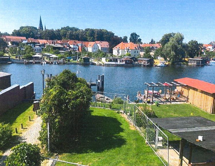 Blick auf einen Garten mit Rasen, Steg und Fluss, dahinter Häuser mit roten Dächern.