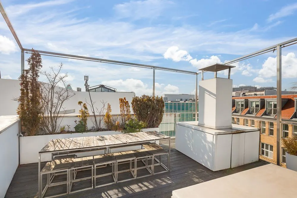 Dachterrasse mit Holztisch, Bänken, Pflanzen und Kamin, umgeben von Stadtgebäuden.