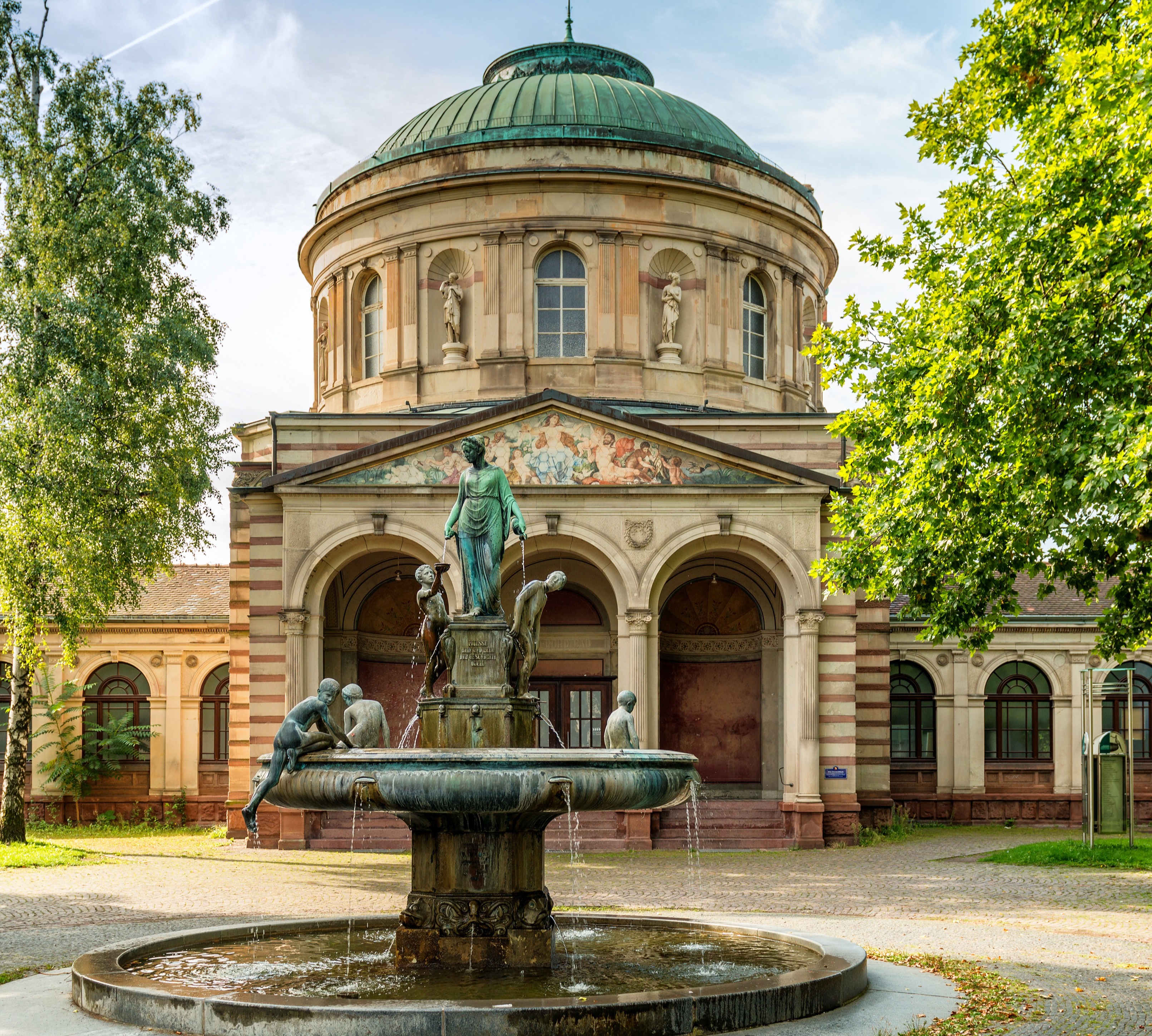 Historisches Gebäude mit Kuppeldach und Brunnen mit Figuren im Vordergrund.