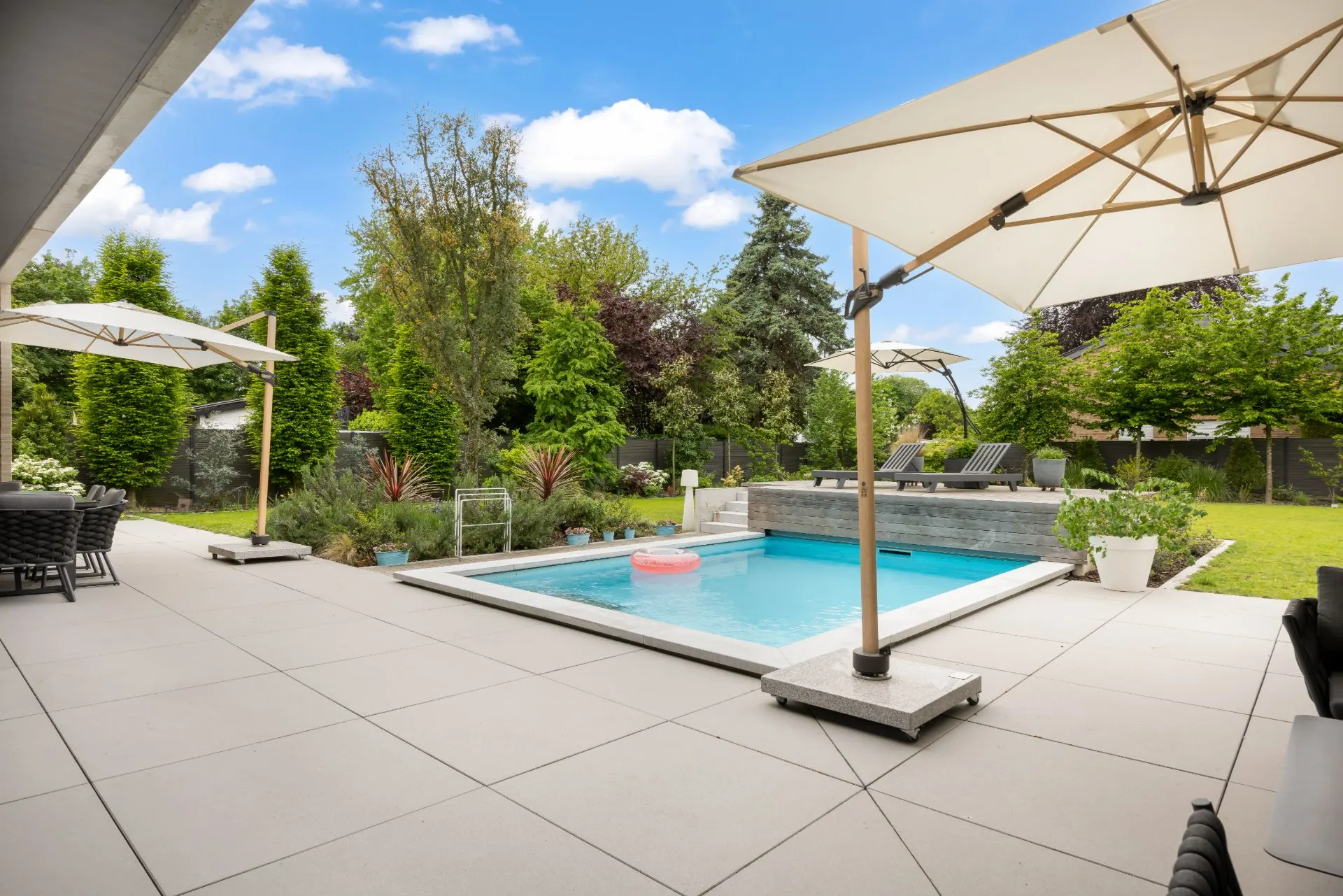 Moderne Terrasse mit Pool, Liegestühlen, Sonnenschirmen und viel Grün im Garten.