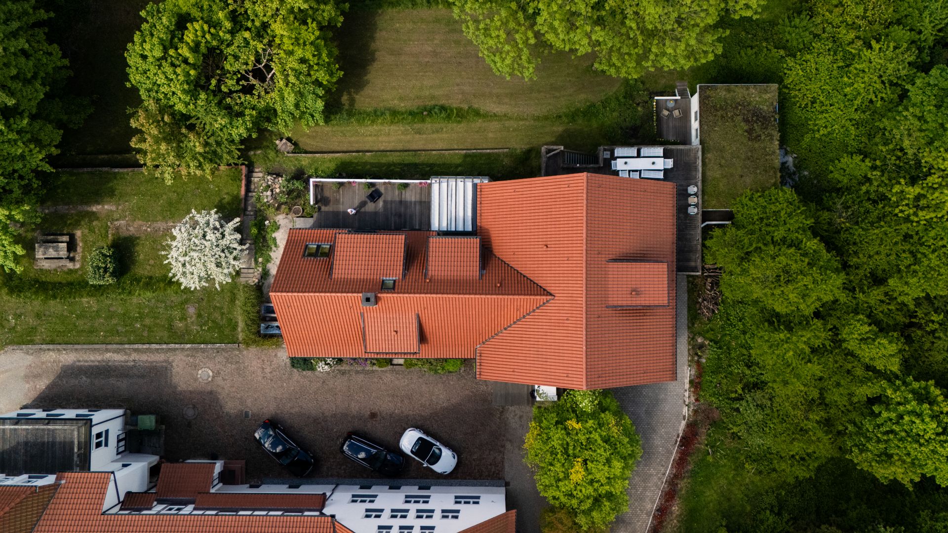 Luftaufnahme eines Hauses mit rotem Dach, umgeben von Bäumen und einer Einfahrt mit geparkten Autos.