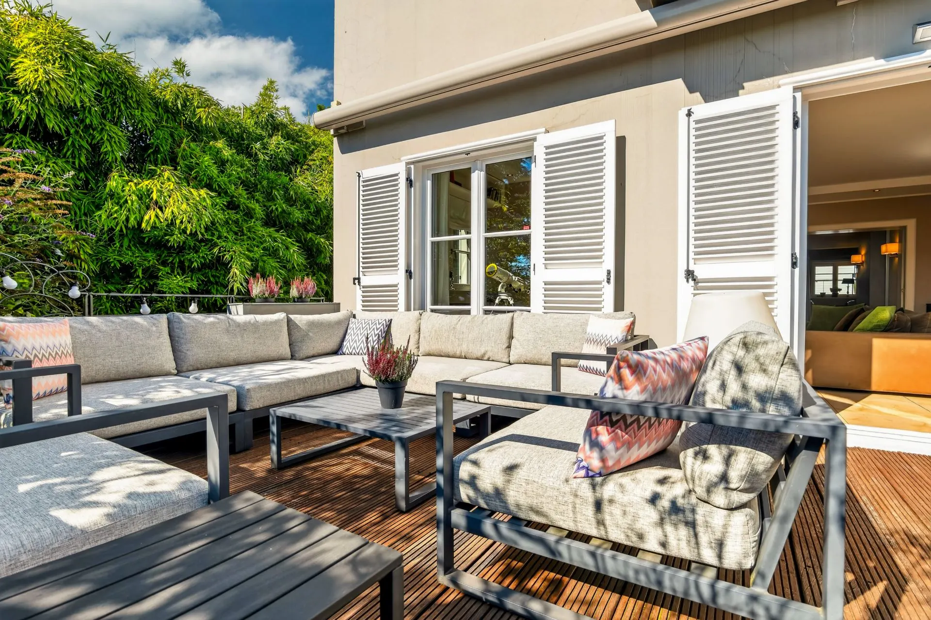 Moderne Terrasse mit Ecksofa, Couchtisch und offenen Fensterläden vor einem Wohnhaus.