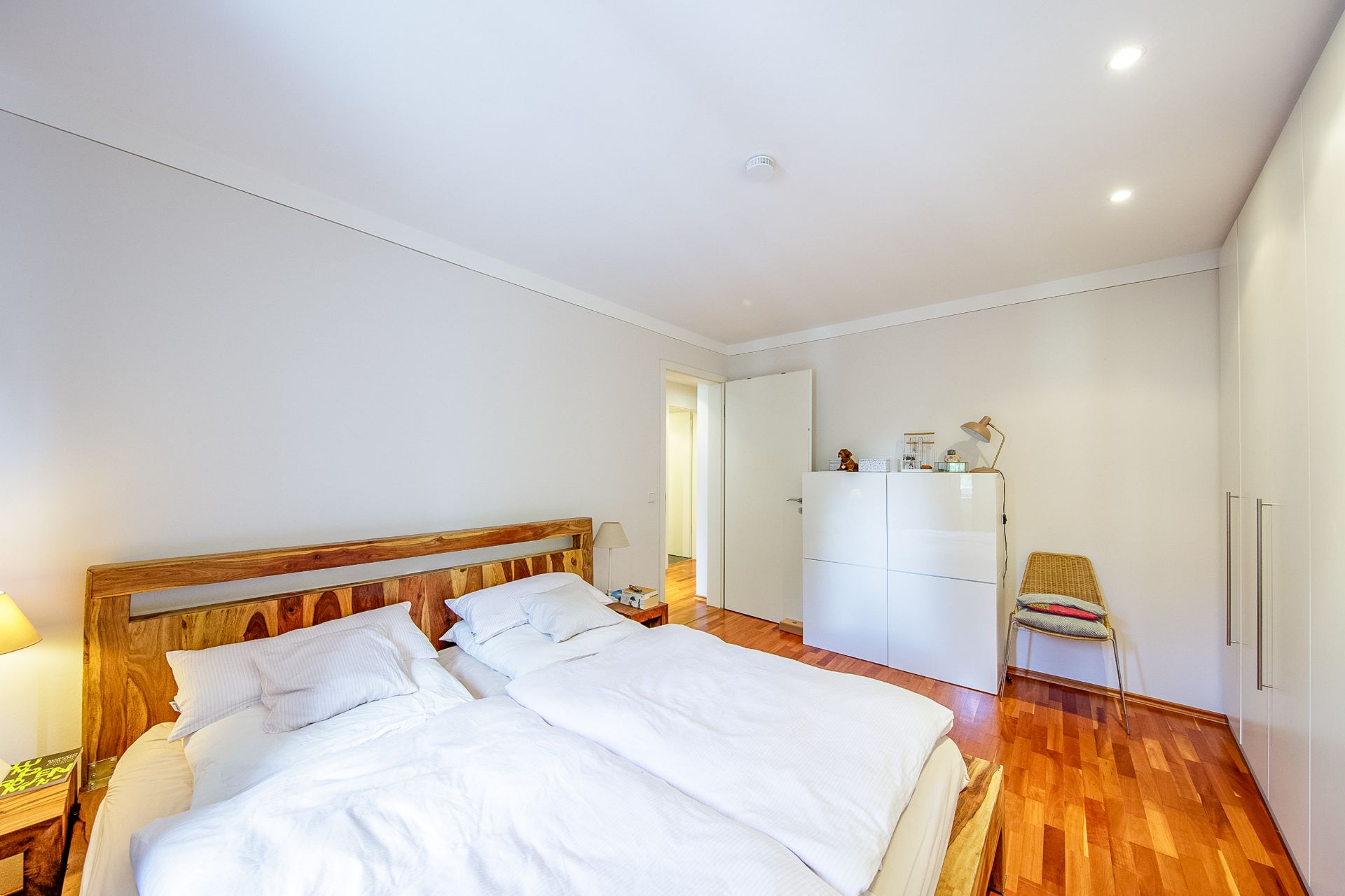 Helles Schlafzimmer mit Doppelbett, Kommode, Stuhl und Holzfußboden.