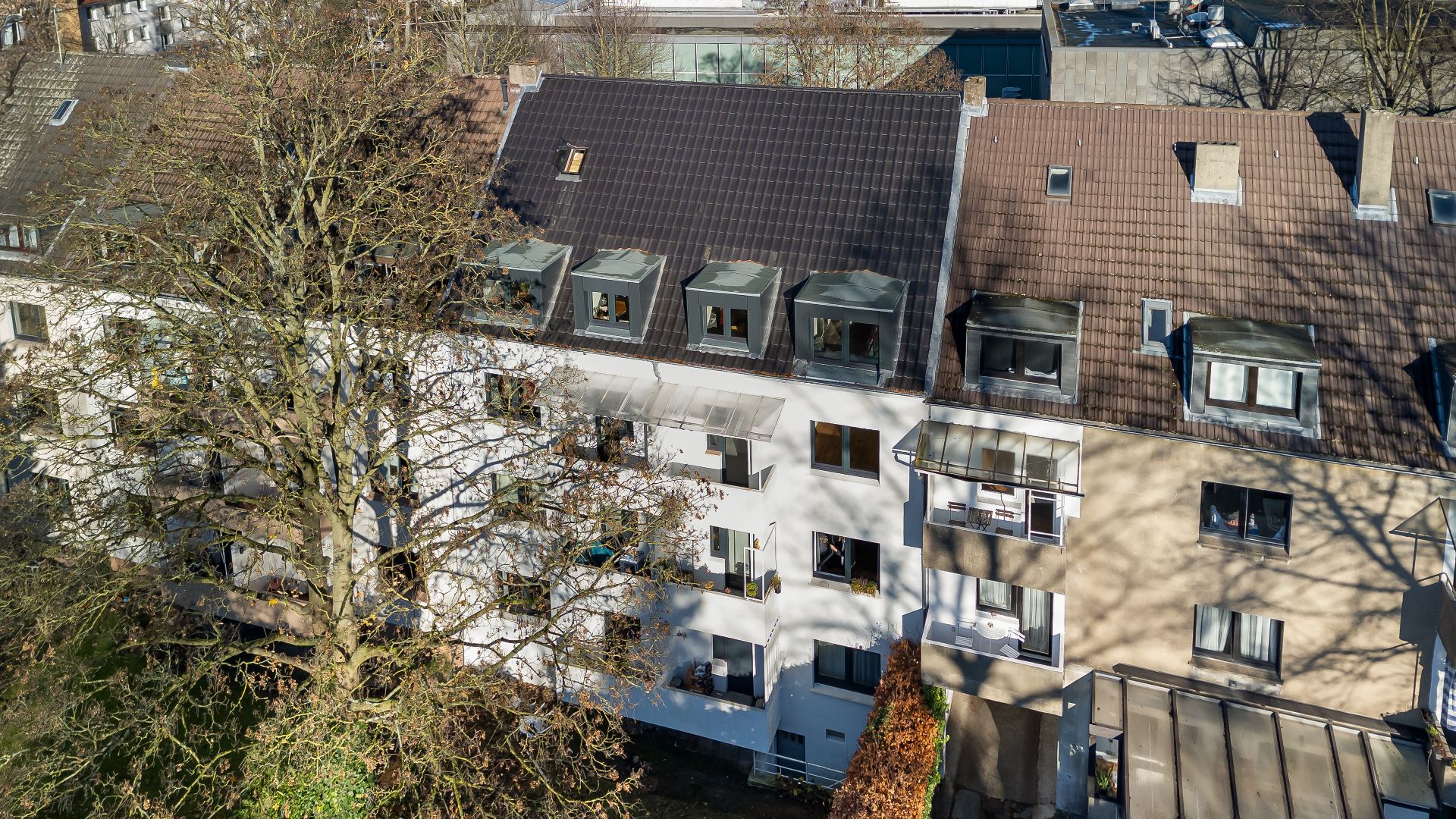 Mehrfamilienhaus mit Balkonen, Dachfenstern und davor stehendem Baum, von oben aufgenommen.