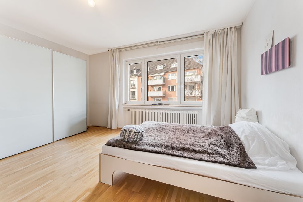 Helles Schlafzimmer mit großem Fenster, Bett und weißem Kleiderschrank auf Holzfußboden.