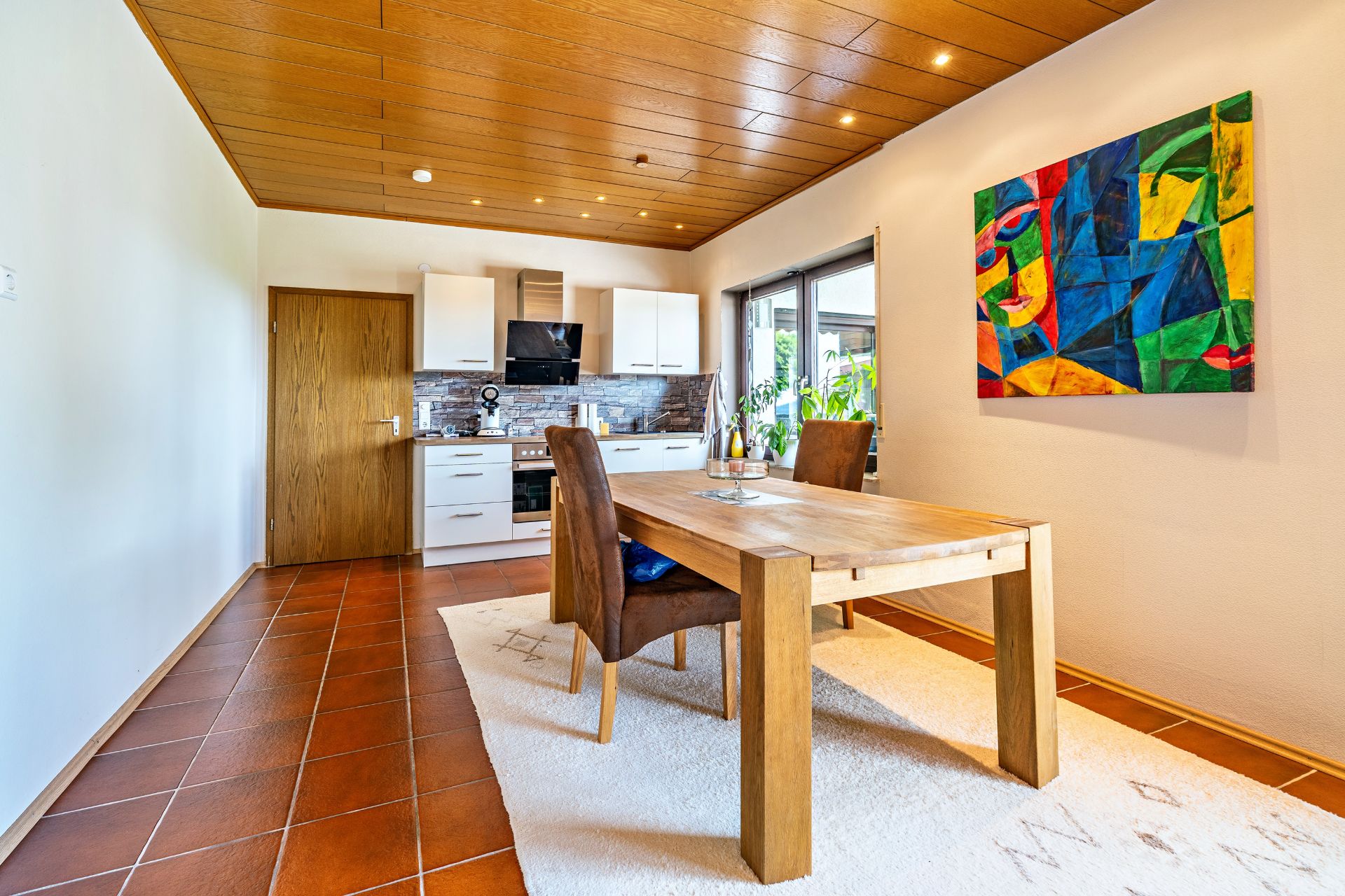 Heller Essbereich mit Holztisch, zwei Stühlen, Teppich, moderner Küche und buntem Bild an der Wand.