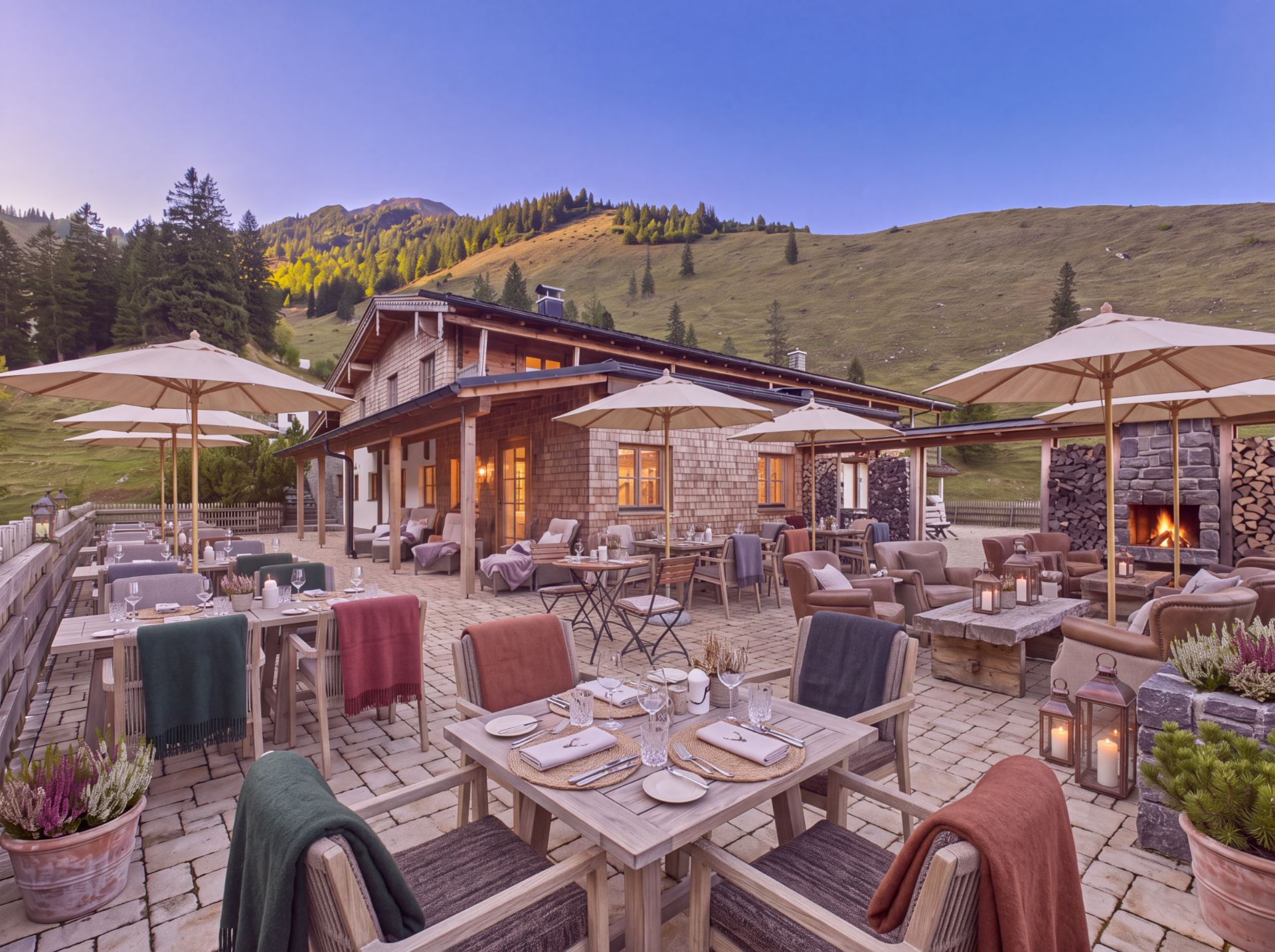 Gemütliche Terrasse mit gedeckten Holztischen, Sonnenschirmen und Blick auf ein Berghaus und grüne Hügel.