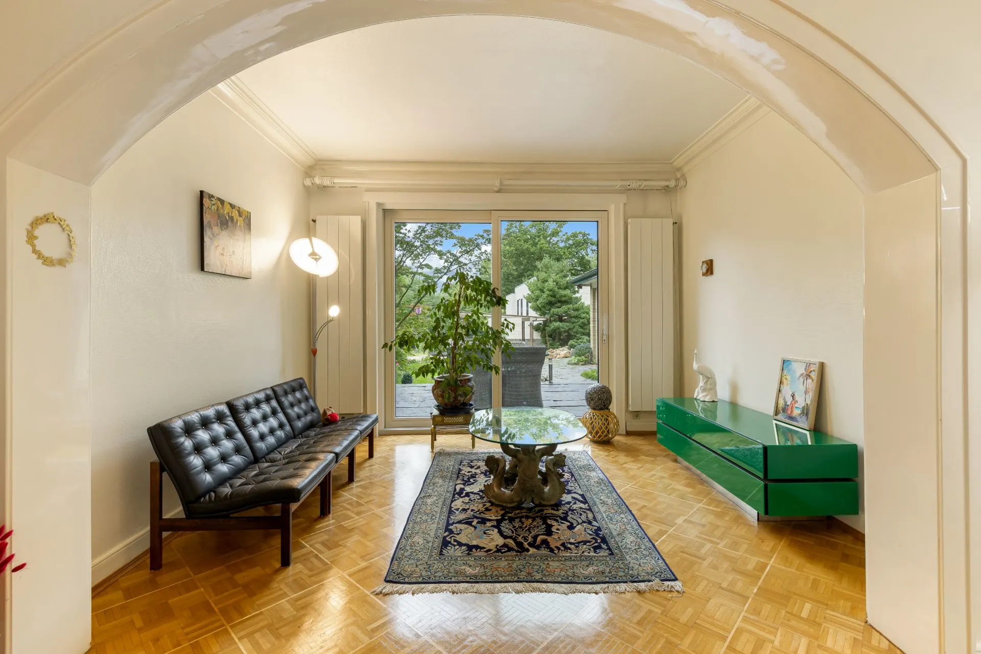 Helles Wohnzimmer mit Sofa, Teppich, Glastisch, Pflanze und großem Fenster zum Garten.
