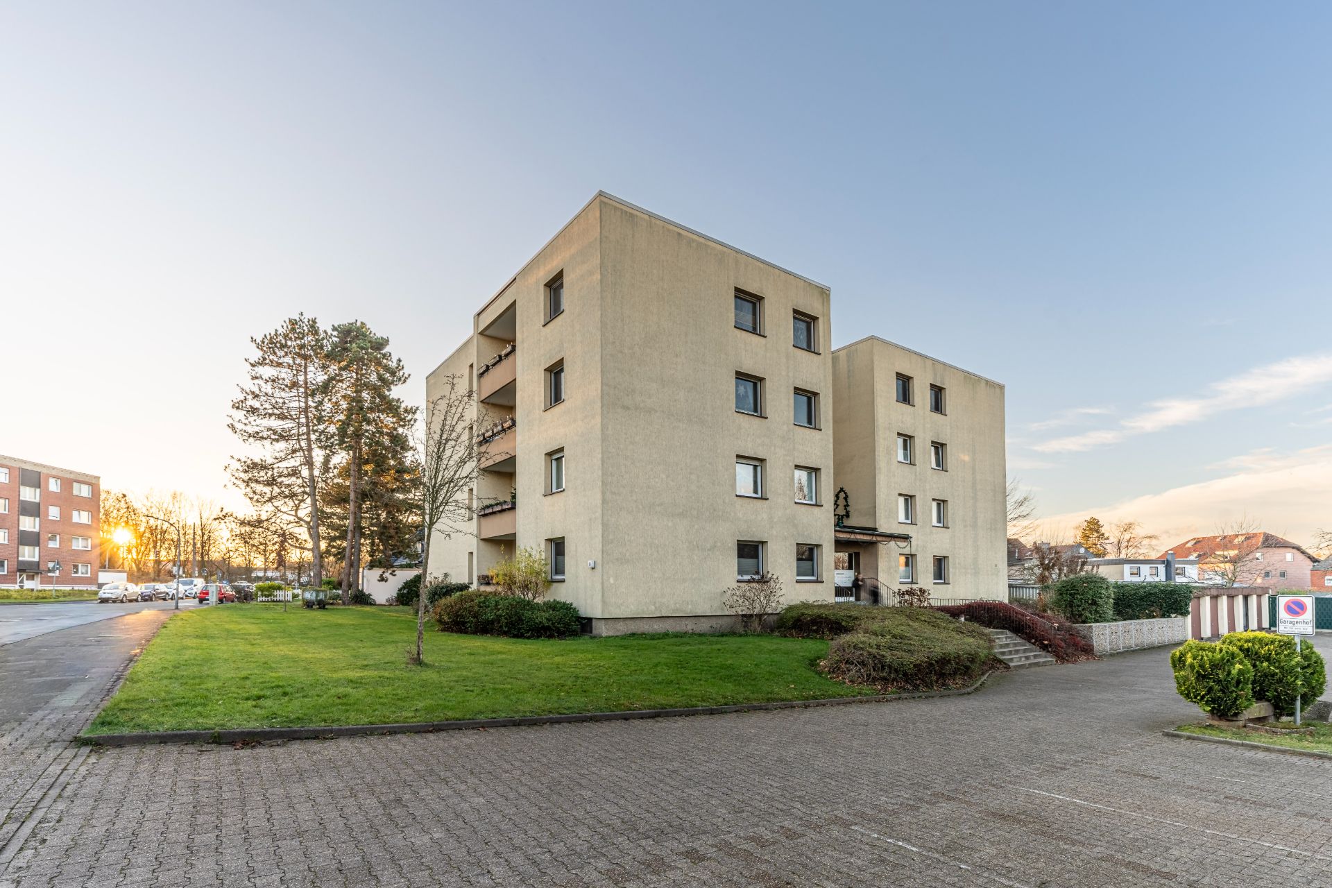 Vierstöckiges Wohngebäude mit Balkonen und gepflegtem Vorgarten bei Sonnenuntergang.