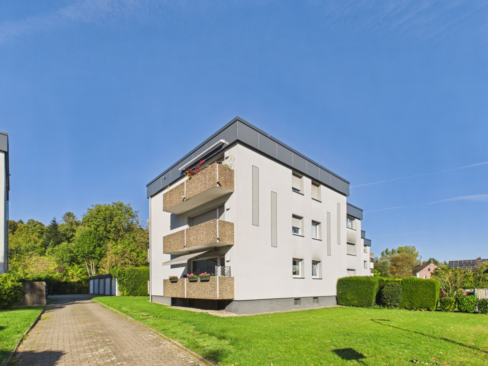 Modernes Wohnhaus mit mehreren Balkonen und grünem Gartenbereich bei Sonnenschein.