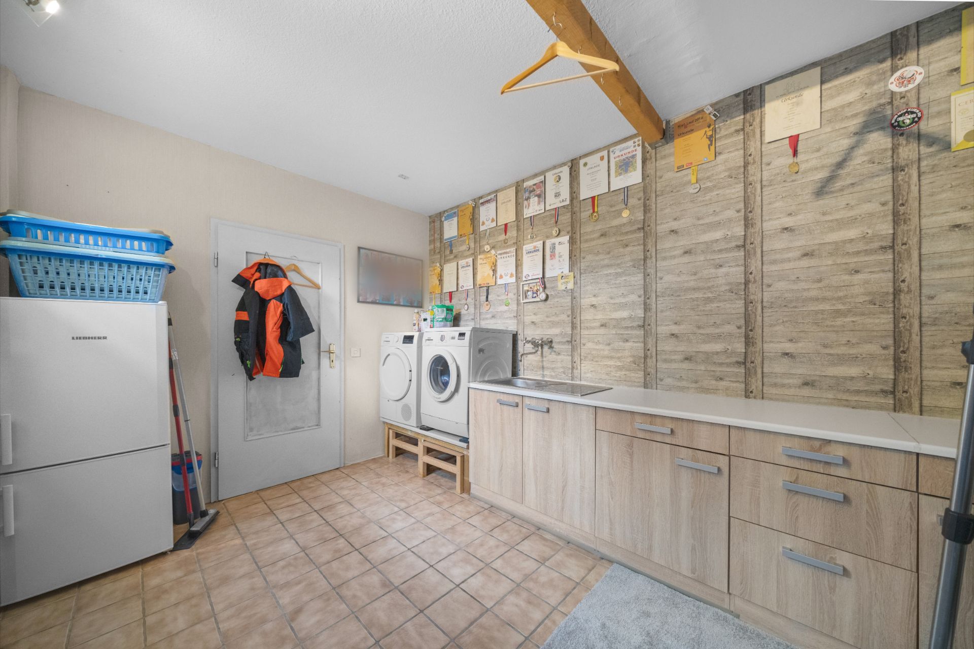 Hauswirtschaftsraum mit Waschmaschine, Trockner, Kühlschrank, Schränken und Urkunden an der Wand.