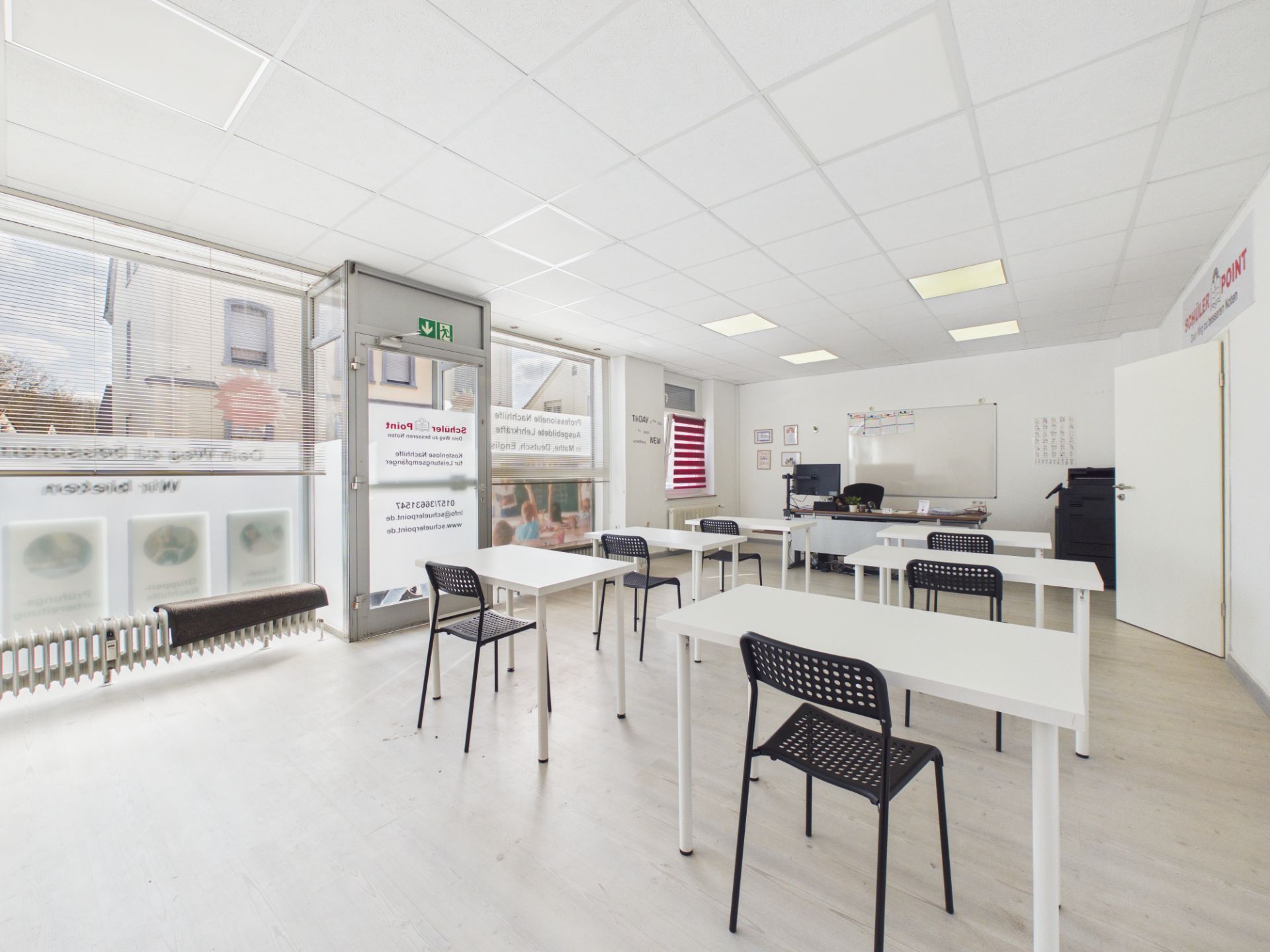 Heller Raum mit mehreren Tischen und Stühlen, großem Fenster und Whiteboard an der Wand.