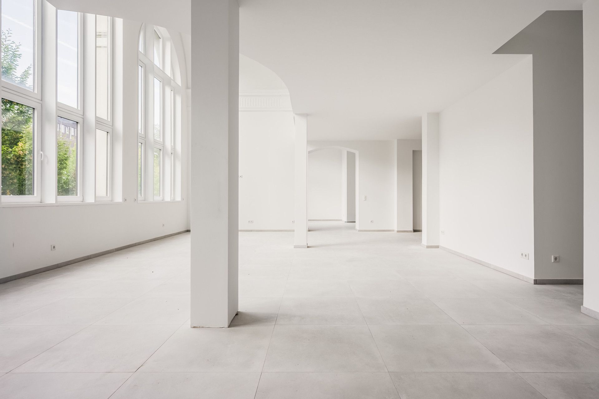 Großer, leerer Wohnraum mit weißen Wänden, Säulen und großen Fenstern auf der linken Seite.