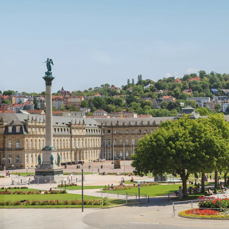 Großer Platz mit hoher Säule, historischen Gebäuden, Bäumen und Blumenbeeten bei sonnigem Wetter.