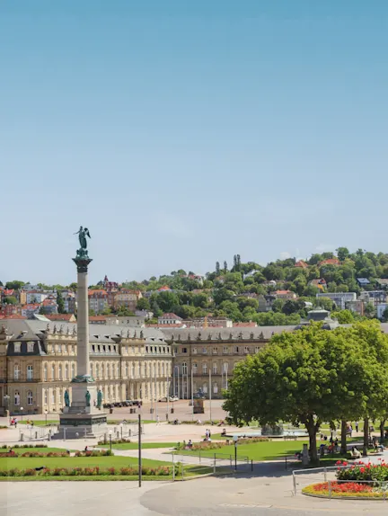 Großer Platz mit hoher Säule, historischen Gebäuden, Bäumen und Blumenbeeten bei sonnigem Wetter.