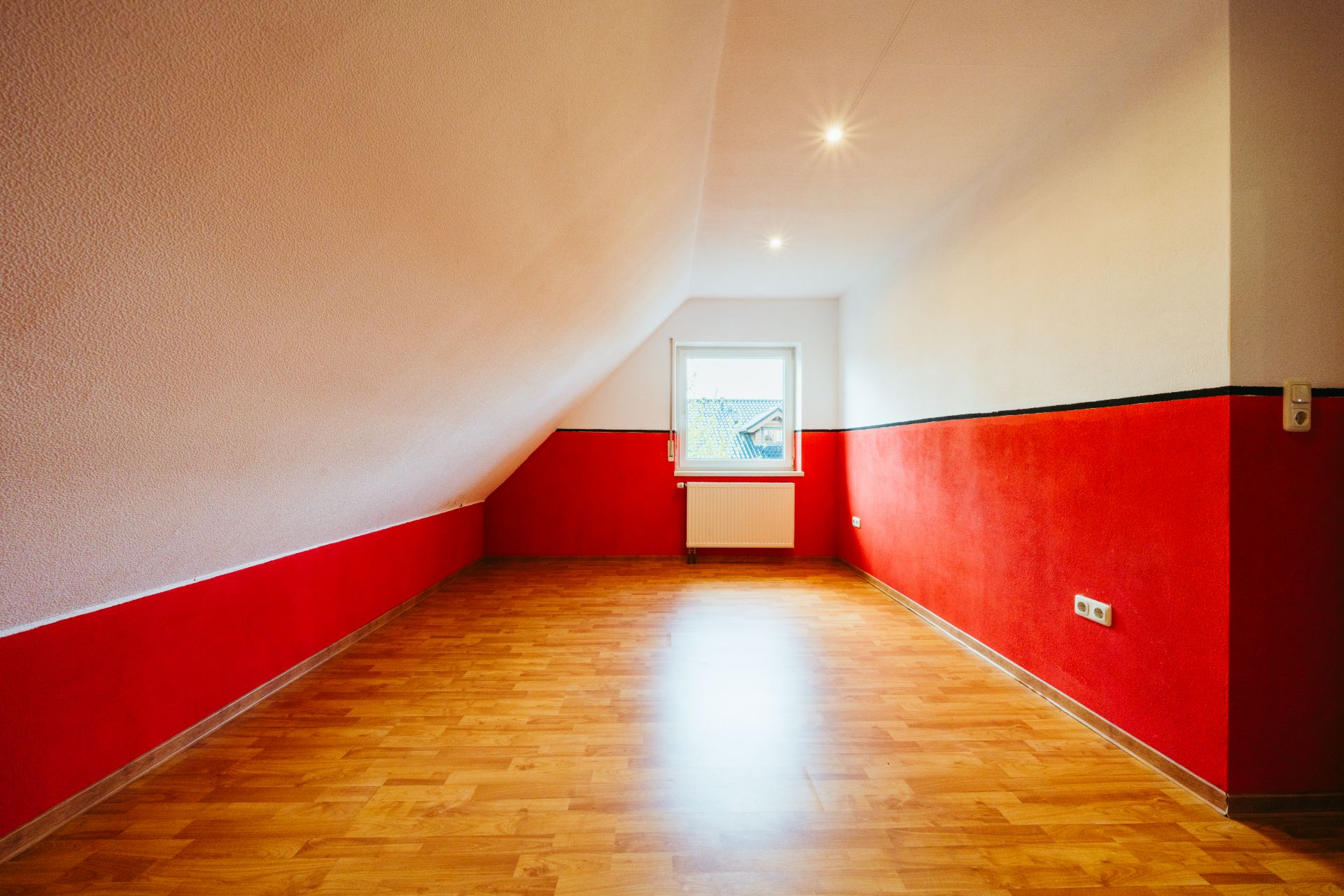 Leeres Zimmer mit schräger Decke, roter Wand, Holzfußboden und Fenster mit Heizkörper.