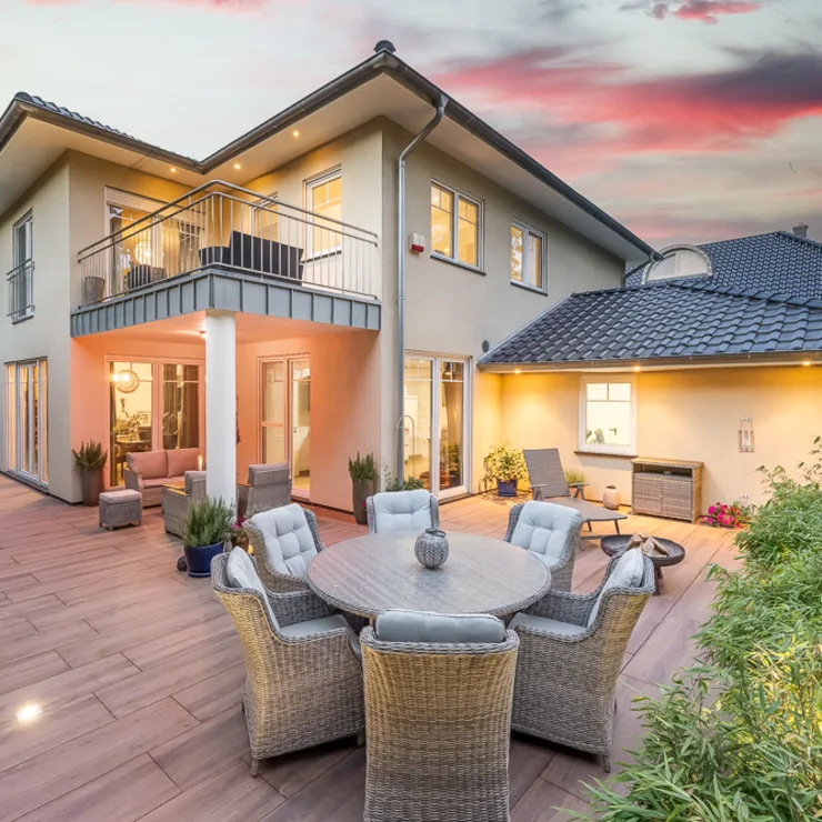 Moderne Terrasse mit Essbereich und gemütlicher Sitzecke vor einem zweistöckigen Wohnhaus bei Sonnenuntergang.
