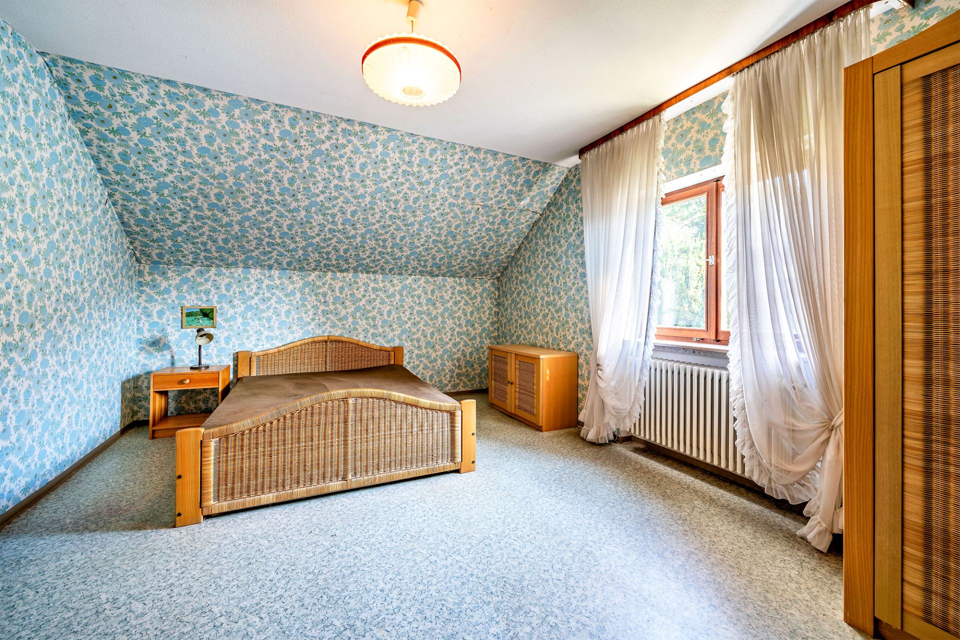 Schlafzimmer mit Holzbett, Nachttisch, Kommode und Fenster mit weißen Vorhängen.