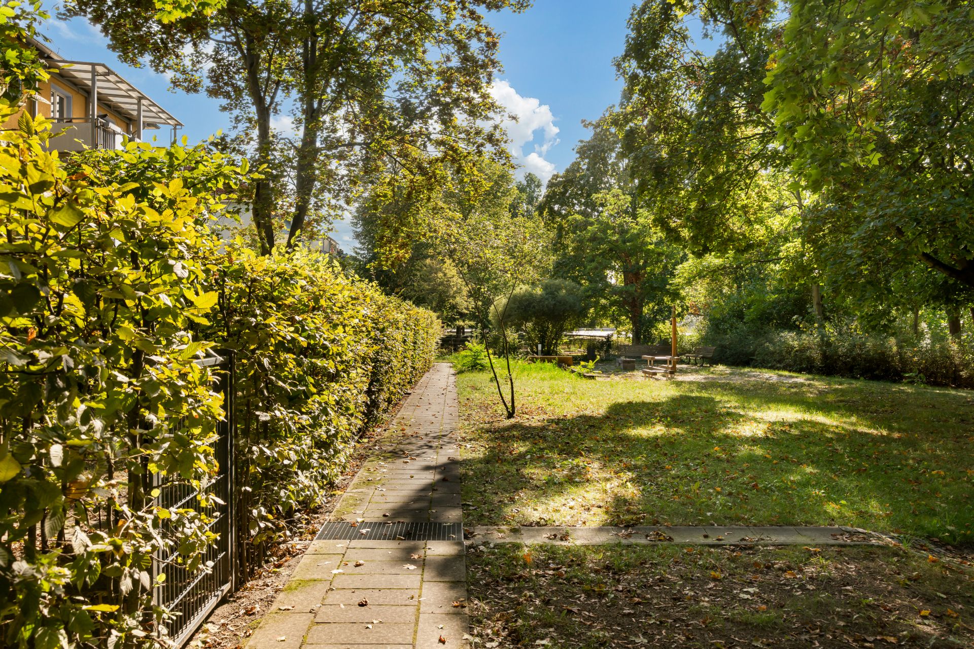 Garten mit Gehweg, Rasenfläche, Bäumen und Bänken im Grünen bei Sonnenschein.