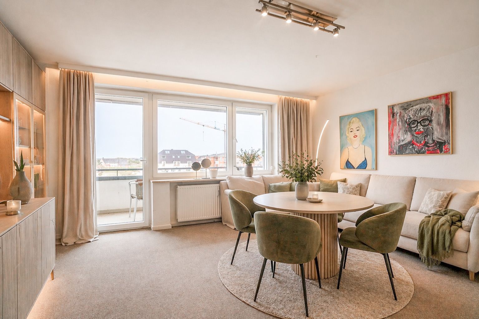 Helles Wohnzimmer mit Esstisch, grünen Stühlen, Sofa und großen Fenstern mit Vorhängen.