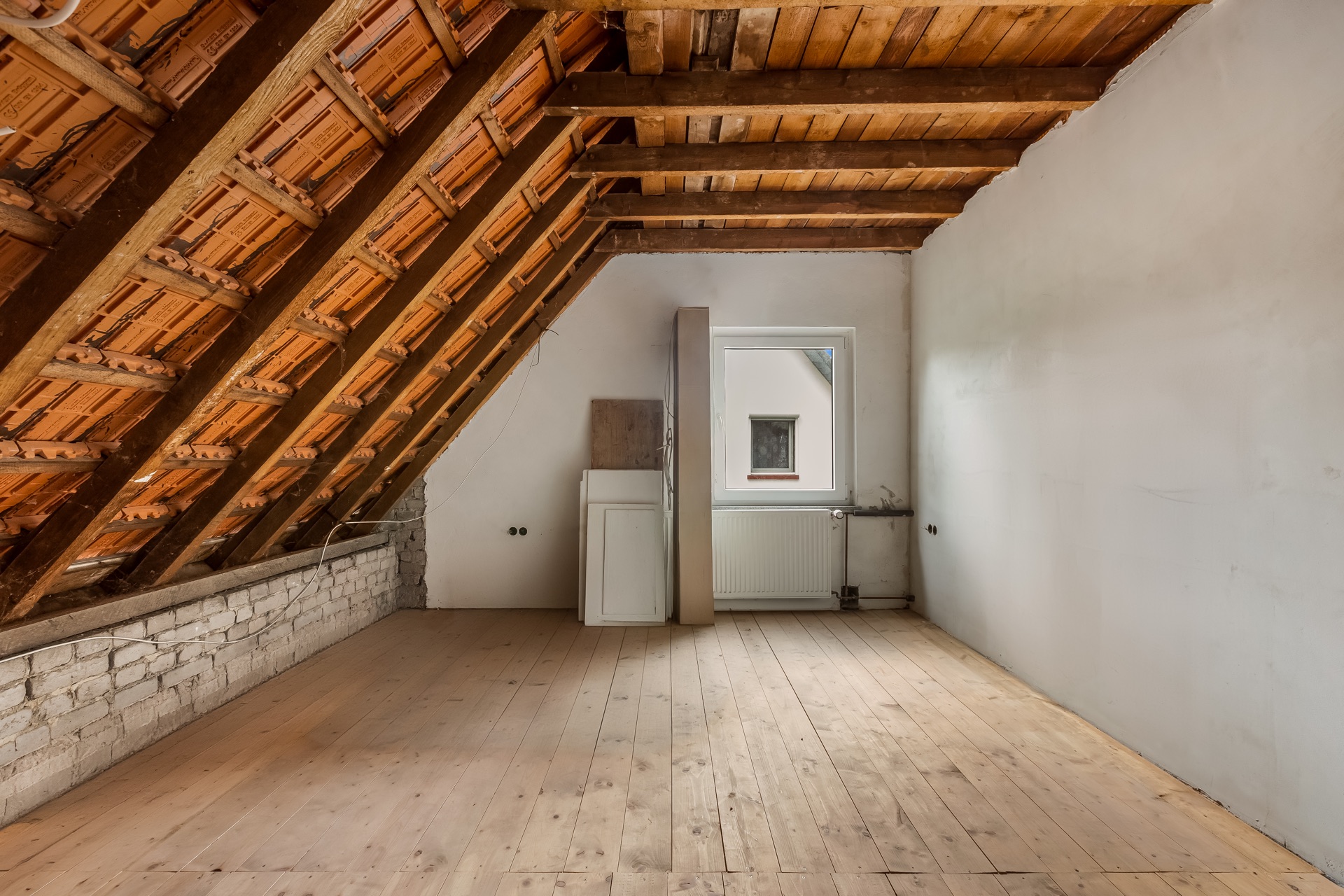 Unoffener Dachboden mit sichtbaren Dachbalken, Holzfußboden, Fenster und Heizkörper.